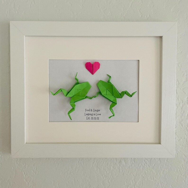 May include: A white framed wall art piece featuring two green origami frogs holding hands beneath a small pink paper heart. Below the frogs, the text reads: Fred & Ginger, Leaping in Love, Est. 01.01.01. The artwork is set against a plain white background.