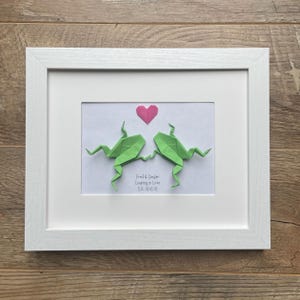 May include: A framed artwork featuring two green origami frogs leaping towards a pink heart. The artwork includes the text "Fred & Ginger Leaping in Love Est. 01 01 01" in a white frame.
