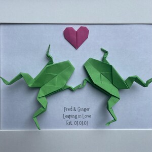 May include: A framed artwork featuring two green origami frogs facing each other with a pink origami heart above them. The text reads: Fred & Ginger, Leaping in Love, Est. 01.01.01. The frogs are positioned on a white background within a white frame.
