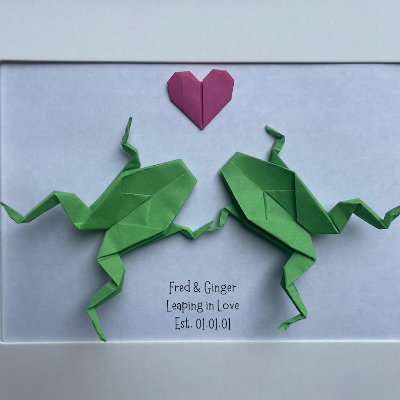 May include: A framed artwork featuring two green origami frogs facing each other with a pink origami heart above them. The text reads: Fred & Ginger, Leaping in Love, Est. 01.01.01. The frogs are positioned on a white background within a white frame.