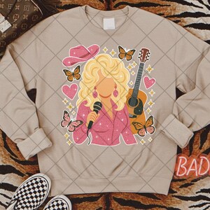 May include: Beige sweatshirt featuring a graphic of a blonde woman in a pink outfit holding a microphone, with a guitar, butterflies, and hearts. A pink cowboy hat is also featured. The word "BAD" is visible on the bottom right.