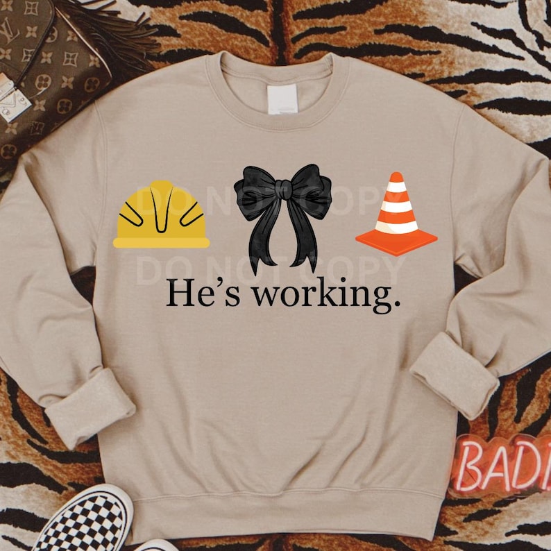 May include: A tan sweatshirt with a yellow hard hat, a black bow, and an orange traffic cone. The text "He's working." is printed below the icons.