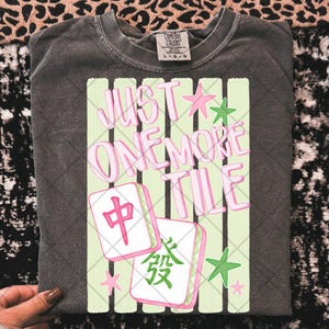 May include: A charcoal grey t-shirt featuring a graphic print with the text JUST ONE MORE TILE in pink and white. The design includes two mahjong tiles with red and green characters, surrounded by small pink and green star shapes on a light green striped background.