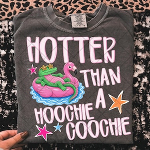 May include: Dark grey Comfort Colors t-shirt with the text "HOTTER THAN A HOOCHIE COOCHIE" in pink and white. The graphic features an alligator wearing sunglasses and a flamingo on a pink float. The shirt is a size L-G-G.