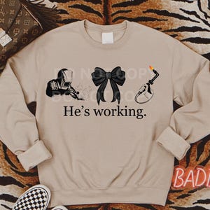 May include: A tan sweatshirt with a black graphic of a welder, a bow, and a welding torch. The text "He's working." is printed below the graphic.