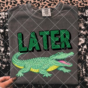 May include: Dark gray t-shirt with the word "LATER" in green block letters above a cartoon alligator. The alligator is green with a yellow belly and pink mouth. The shirt is a Comfort Colors brand.