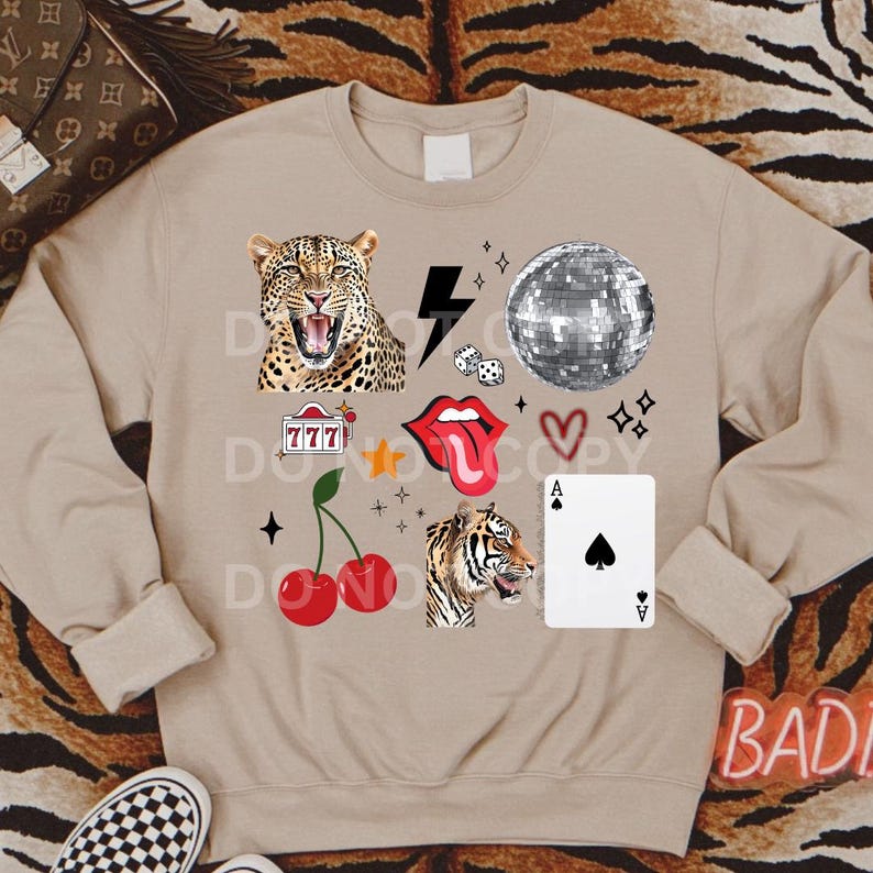 May include: A tan sweatshirt featuring a collection of colourful patches, including a leopard, a tiger, a cherry, a disco ball, a playing card, a slot machine, a tongue, and a heart. The sweatshirt has the word "BAD" printed on the bottom right corner.