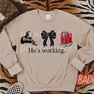 May include: A tan sweatshirt with a black welder, a black bow, and a red welding machine. The text "He's working." is printed below the images.