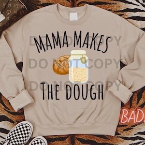 Puede incluir: Una sudadera de color beige con el texto "Mama Makes The Dough" en negro. Hay un dibujo de un tarro de masa y una hogaza de pan.