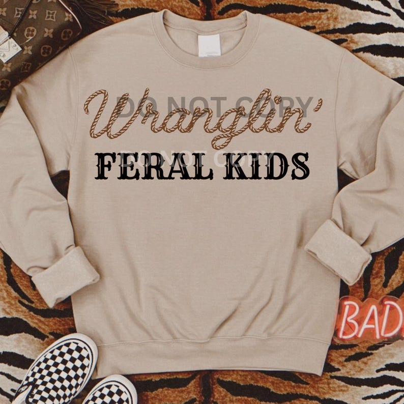 Może przedstawiać: Beżowa bluza z napisem "Wranglin' Feral Kids" w czarnych literach. Tekst jest stylizowany na wz&oacute;r liny.