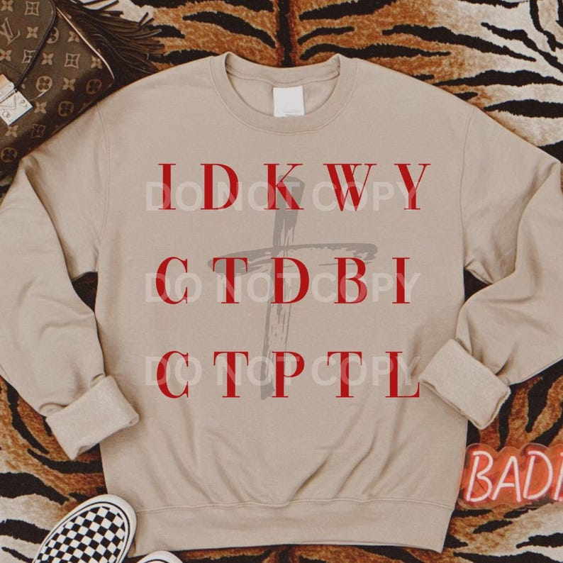 May include: A beige sweatshirt with a red cross and the text "IDK WY CTDBI CTP TL" printed on it.