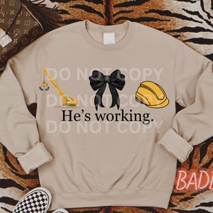 May include: A tan sweatshirt with a yellow crane, a black bow, and a yellow hard hat. The text "He's working." is printed on the sweatshirt.
