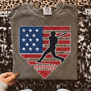 May include: A gray t-shirt with a vintage-style American flag and baseball player graphic. The graphic is in red, white, and blue and features a baseball player in a batting stance. The text "COMFORT COLORS" and "L-G-O" is visible on the shirt.