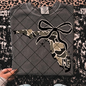 Puede incluir: Una camiseta gris con un contorno de camuflaje del estado de Florida con un lazo negro en la parte superior y las letras "FL" dentro del contorno.