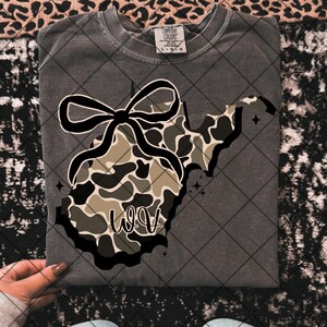 May include: Dark grey t-shirt with a West Virginia outline, camouflage pattern, and a black bow. The letters "WV" are in a script font. The shirt is a Comfort Colors brand.