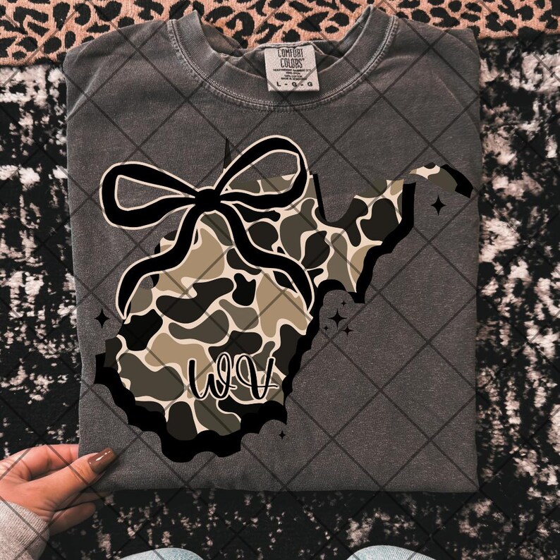 May include: Dark grey t-shirt with a West Virginia outline, camouflage pattern, and a black bow. The letters "WV" are in a script font. The shirt is a Comfort Colors brand.