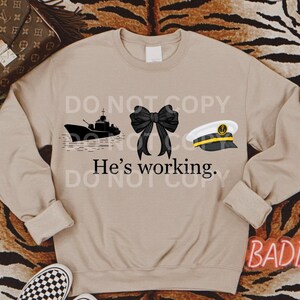 May include: A tan sweatshirt with a black ship, a black bow, and a black and white sailor hat. The text "He's working." is printed on the sweatshirt.
