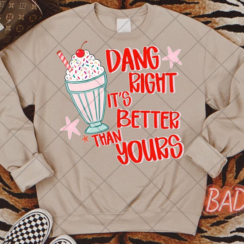 May include: A beige crewneck sweatshirt featuring a graphic of a milkshake with a cherry on top, a red and white striped straw, and colorful sprinkles. The text reads DANG RIGHT IT'S BETTER THAN YOURS in red letters, accented by two small pink stars.