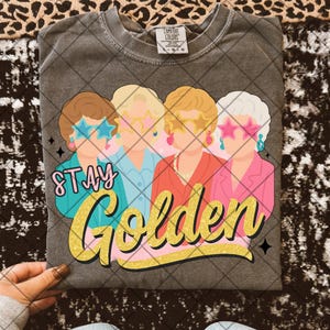 Puede incluir: Una camiseta gris con un gráfico de cuatro mujeres con gafas de sol y el texto "Stay Golden".