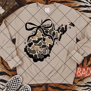 May include: A beige sweatshirt with a camouflage design in the shape of West Virginia, a black bow, and the word "WOW". The design includes black and white stars.
