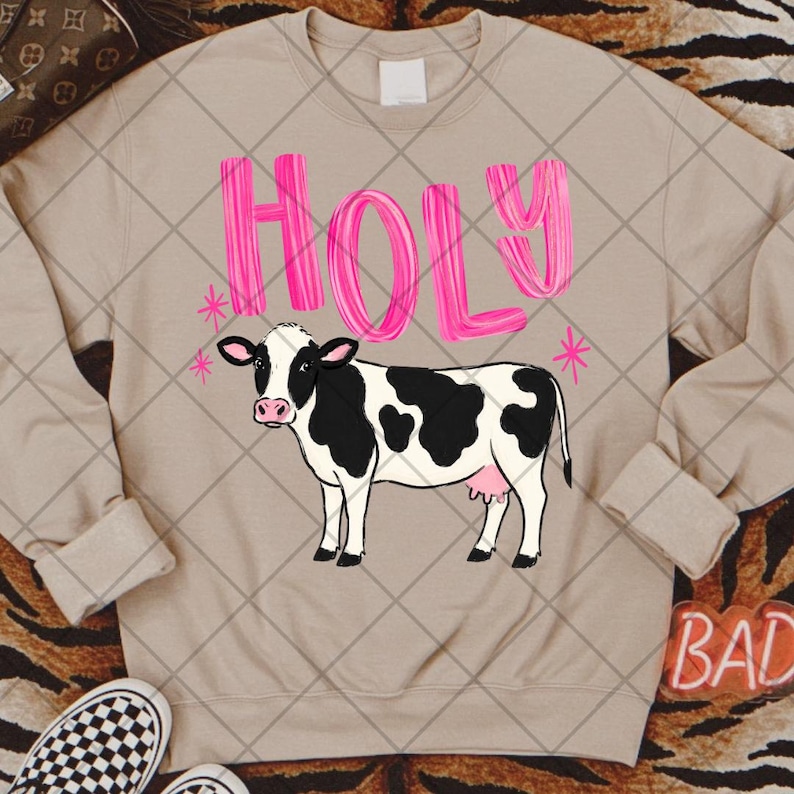 Puede incluir: Una sudadera beige de manga larga con el gr&aacute;fico de una vaca blanca y negra. Sobre la vaca, la palabra HOLY est&aacute; impresa en letras grandes, texturizadas y de color rosa brillante. Peque&ntilde;os iconos de estrellas rosas aparecen a ambos lados.