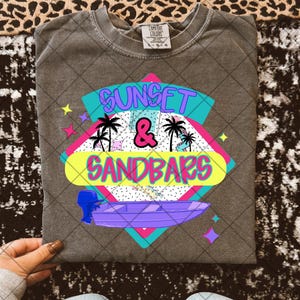May include: A gray t-shirt with a retro-style graphic design featuring the text "Sunset & Sandbars" in pink, blue, and yellow. The graphic also includes palm trees, a boat, and stars.