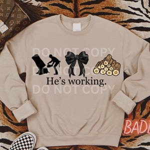 May include: A tan sweatshirt with a black silhouette of a person chopping wood, a black bow, and a pile of wood logs. The text "He's working." is printed below the images.