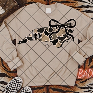 以下が含まれることがあります： ベージュのトレーナーに、カモフラージュ柄のノースカロライナ州のアウトラインが描かれており、その上に黒いリボンが置かれています。アウトラインの中には「NC」の文字があります。