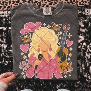 May include: Dark gray Comfort Colors t-shirt featuring a graphic of a blonde woman with a microphone, a pink cowboy hat, butterflies, a guitar, and hearts. The woman is wearing a pink outfit and earrings. The shirt is a size L-G-G.
