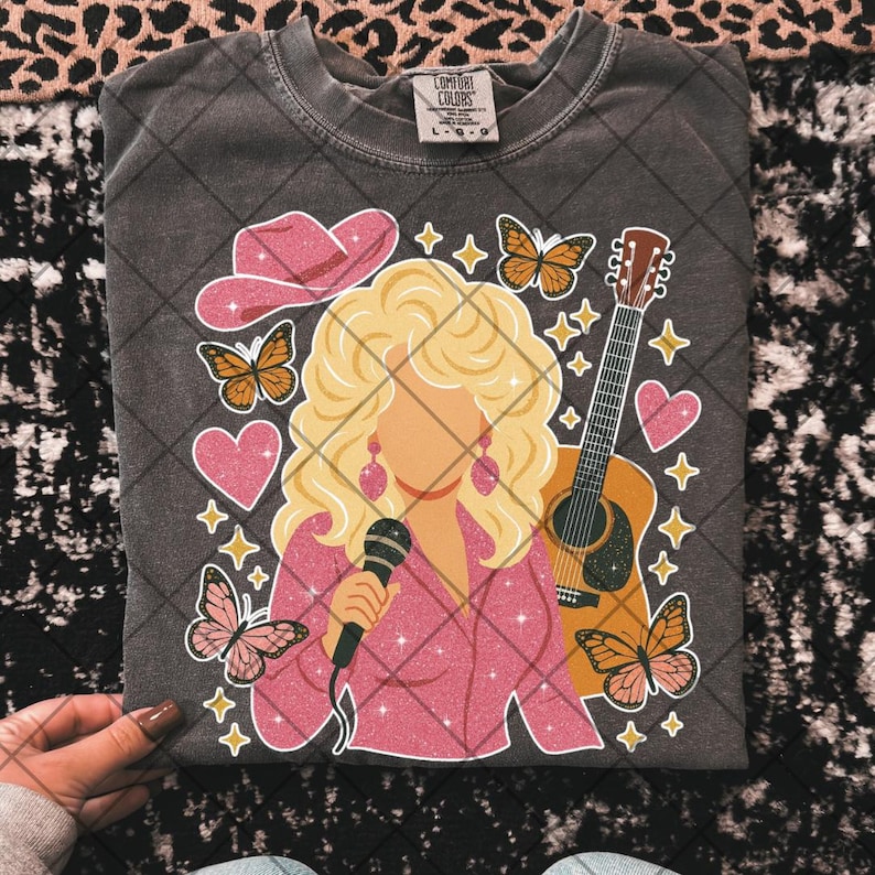 May include: Dark gray Comfort Colors t-shirt featuring a graphic of a blonde woman with a microphone, a pink cowboy hat, butterflies, a guitar, and hearts. The woman is wearing a pink outfit and earrings. The shirt is a size L-G-G.