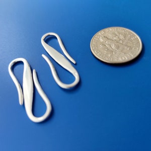 Puede incluir: Dos cierres de gancho plateados, curvados y pulidos. Se muestran sobre un fondo azul, junto a una moneda para comparar el tamaño. Estos cierres son probablemente para joyería u otros proyectos de artesanía.