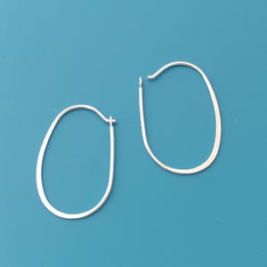 Op de afbeelding: Een paar zilverkleurige ovale oorringen. De oorbellen hebben een eenvoudig, minimalistisch ontwerp met een gladde, gepolijste afwerking. De ringen zijn licht afgeplat en hebben een haaksluiting. De achtergrond is effen blauwgroen.