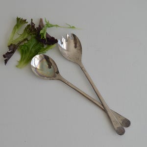 May include: Two silver salad servers, a spoon and a fork, are crossed on a white surface. A small pile of fresh green and purple lettuce is next to the servers. The spoon has a rounded bowl, and the fork has three tines.
