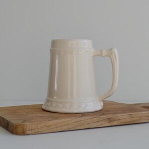 Puede incluir: Una jarra de cerveza de cerámica color crema con asa, apoyada sobre una tabla de cortar de madera. La jarra tiene una forma clásica con una banda decorativa cerca de la base y la parte superior. La tabla de cortar es rectangular con asa.