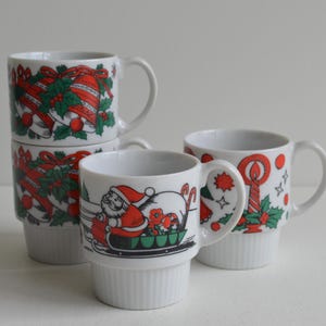 Könnte beinhalten: Weiße Keramikbecher mit weihnachtlichen Designs. Ein Becher zeigt den Weihnachtsmann im Schlitten, andere Glocken, Stechpalmen und Zuckerstangen. Die Becher haben einen gerippten Boden und einen gebogenen Griff, ideal für Feiertagsgetränke.