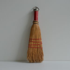 May include: A small hand broom featuring a natural tan straw bristle head secured with red twine. The handle is painted red and topped with a silver-colored metal cap that includes a small loop for hanging the item vertically.