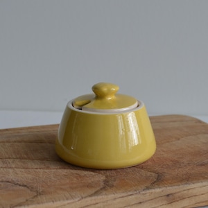 May include: A small, yellow ceramic sugar bowl with a lid and a small handle. The bowl is sitting on a wooden cutting board. The background is a plain, light gray color.