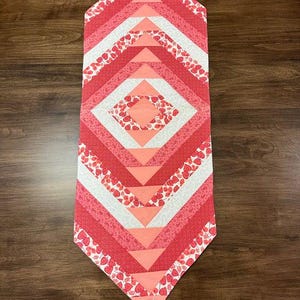 Peut inclure: Chemin de table matelassé avec un motif géométrique. Le chemin de table présente un motif de losanges dans les tons de rouge, de rose et de blanc. Certaines sections ont un imprimé fraise. Le chemin de table mesure environ 102 cm de long.