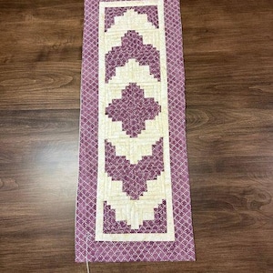 Peut inclure: Chemin de table matelassé rectangulaire avec un motif géométrique. Le chemin de table présente un motif central de blocs violets et crème, encadré par une bordure à motifs violets et blancs. Le chemin de table mesure environ 30 cm de large et 76 cm de long.