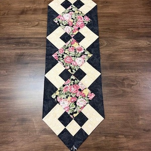 Peut inclure: Chemin de table matelassé avec un motif de losanges noirs et crème. Le chemin de table présente trois patchs floraux en forme de losange avec des roses roses et jaunes sur fond noir. Le chemin de table est présenté sur une surface en bois.