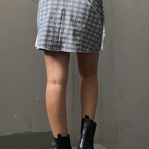 May include: A blue and white plaid mini skirt with white ties on the sides. The skirt is being worn with black platform boots.