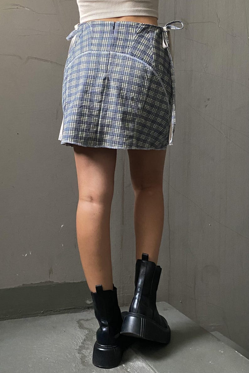 May include: A blue and white plaid mini skirt with white ties on the sides. The skirt is being worn with black platform boots.