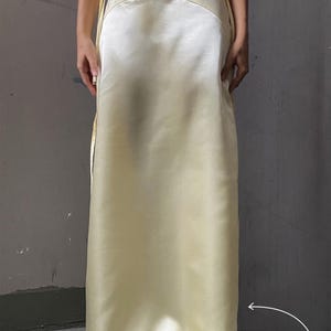 May include: A light yellow satin skirt with a high waist and a straight, flowing silhouette. The skirt is worn by a person standing with their feet together, and the skirt is pulled to the side to show the hemline.