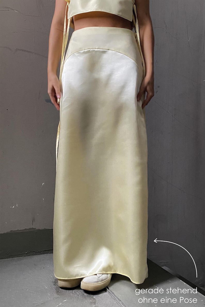 May include: A light yellow satin skirt with a high waist and a straight, flowing silhouette. The skirt is worn by a person standing with their feet together, and the skirt is pulled to the side to show the hemline.