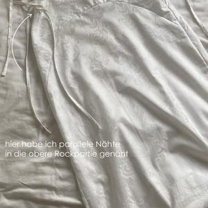 May include: White two-piece outfit with a floral pattern. The top has a halter neck and ties at the back. The skirt has a high waist and ties at the sides. The text "hier habe ich parallele N&auml;hte in die obere Rockpartie gen&auml;ht" is written on the image.