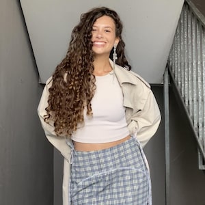 May include: A woman wearing a white crop top, a beige trench coat, and a blue and white plaid skirt. She has long, curly brown hair and is smiling.
