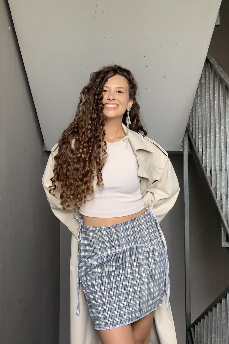 May include: A woman wearing a white crop top, a beige trench coat, and a blue and white plaid skirt. She has long, curly brown hair and is smiling.