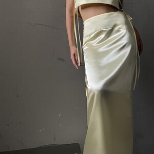 May include: A two-piece outfit in a pale yellow satin fabric. The top is a sleeveless crop top with a square neckline and ties at the sides. The skirt is a long, flowing skirt with a high waist.