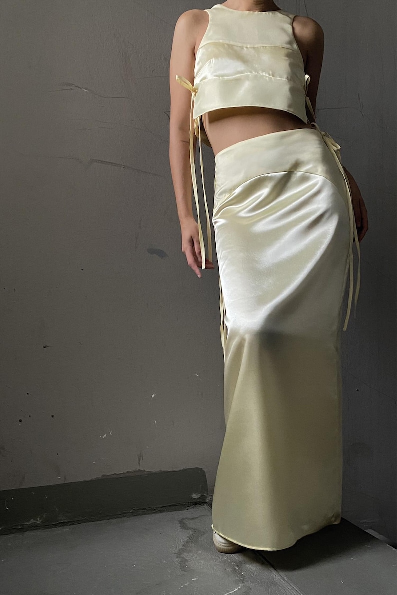 May include: A two-piece outfit in a pale yellow satin fabric. The top is a sleeveless crop top with a square neckline and ties at the sides. The skirt is a long, flowing skirt with a high waist.