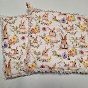 May include: Two quilted potholders with a white background featuring watercolor-style rabbits and floral arrangements in yellow, pink, and purple. The potholders have a frayed edge and a loop for hanging.